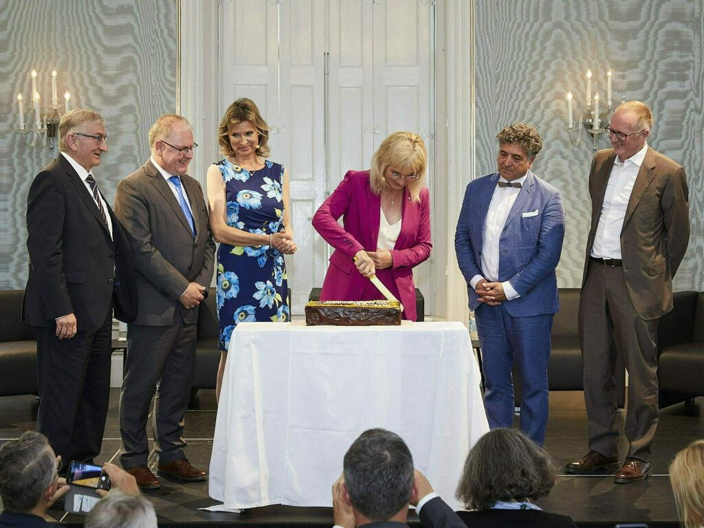 Frau Staatsministerin Scharf schneidet zum 100 jährigen Bestehen der Schwerbehindertenvertretung im Beisein der an der Moderation Teilnehmenden eine Geburtstagstorte an.